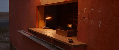 A kiosk serving hatch at nightfall, desk lamp lit, a feature phone on the counter and prices chalked on the wall