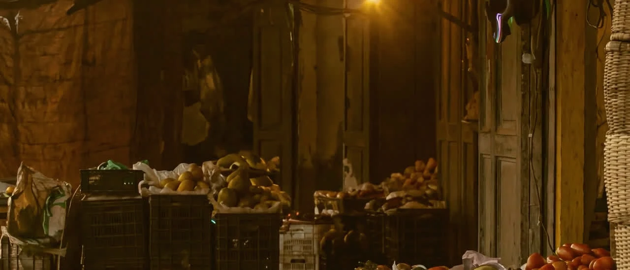 A covered market aisle at dusk seen up close, stalls stacked with produce and one bulb lit