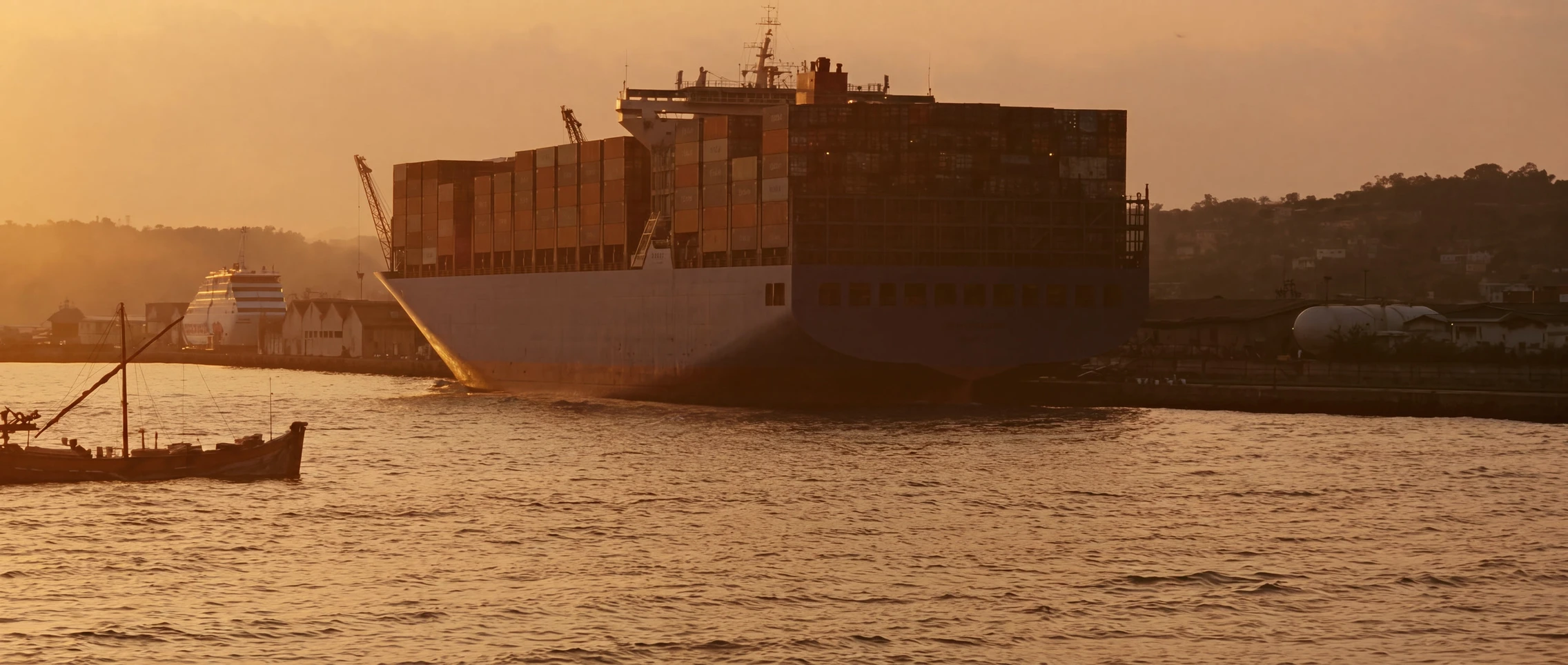 A loaded container ship at the harbour mouth at dusk, a wooden boat crossing in the foreground