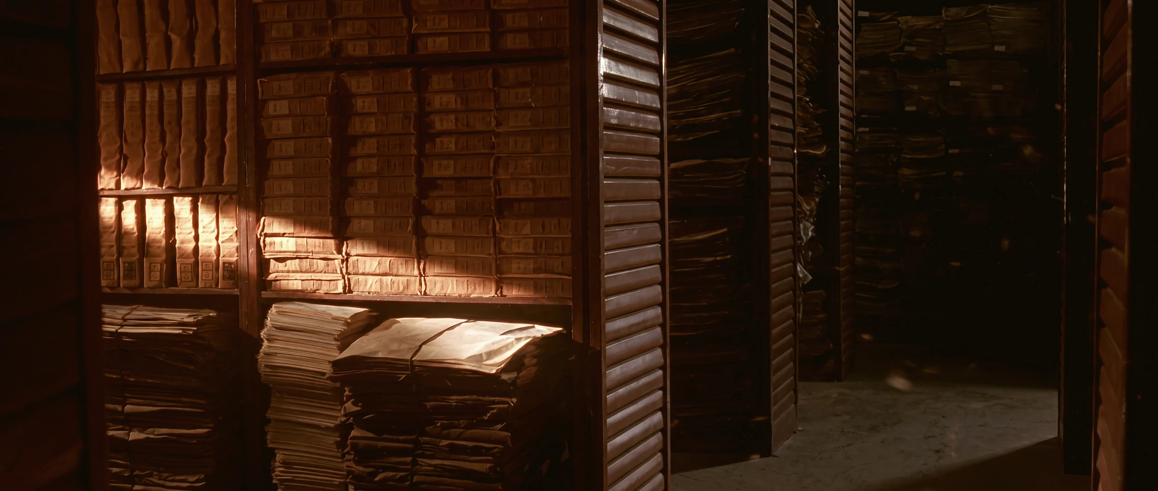 Bundled paper files stacked on the wooden shelves of a records room, one warm shaft of light across them