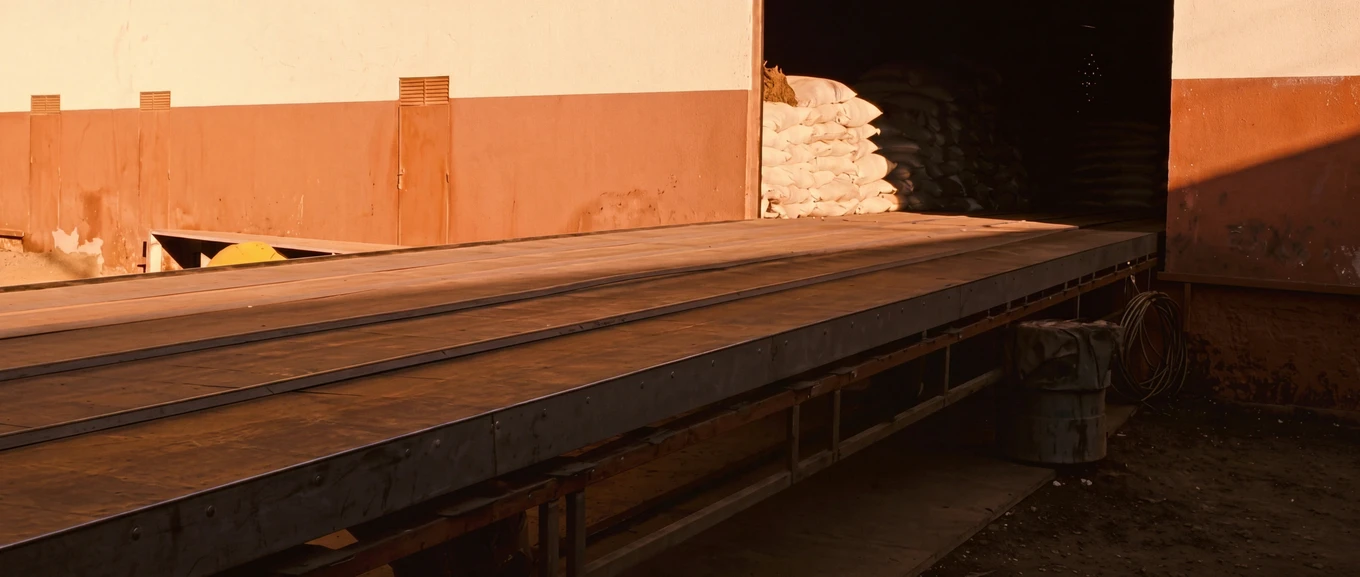 An empty weighbridge platform in evening light, sacks of grain stacked in the warehouse doorway behind it